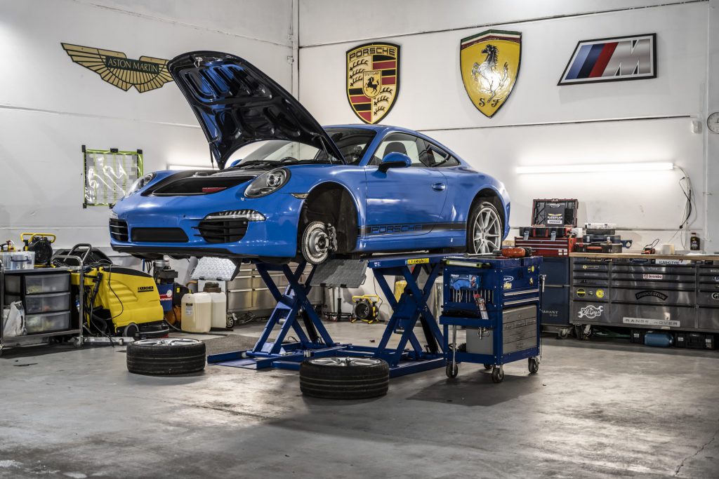 A blue Porsche is lifted in a Garage with the bonnet open and front wheels removed. In the background is lots of tools and manufactere badges are displayed on the walls